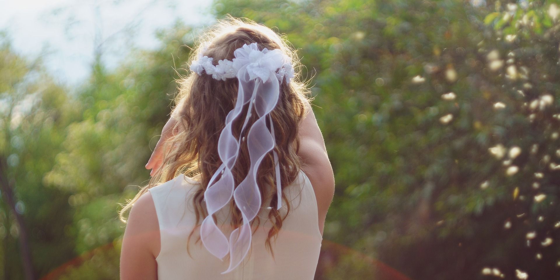 Na obrazie widzimy kobietę z długimi włosami, ukazaną z tyłu. Jej głowę zdobi biały ozdobny wianek, który zawiera wstążki. Scena rozgrywa się na zewnątrz, w otoczeniu zieleni i naturalnego światła. 